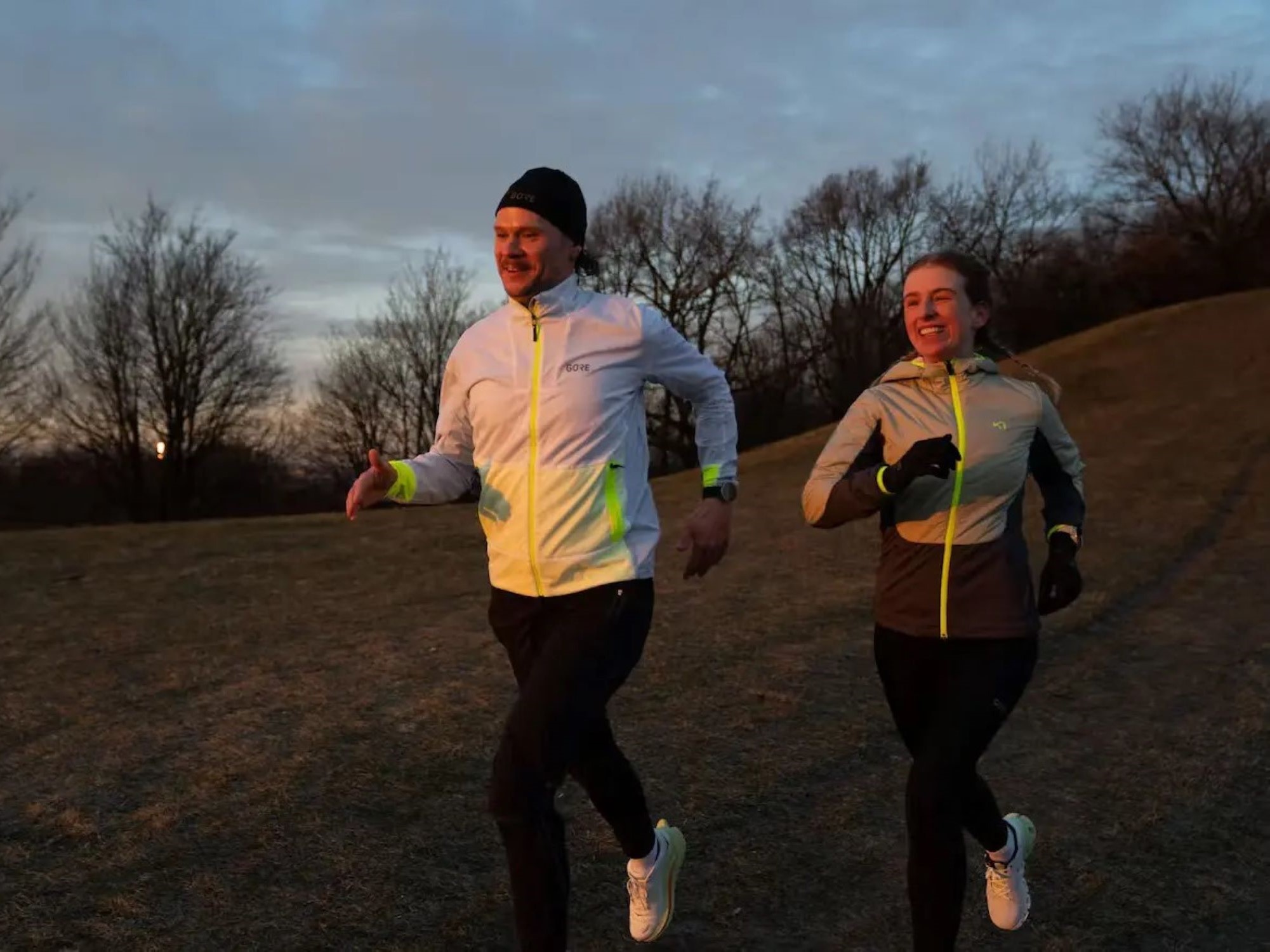 Bild Zwei Personen joggen. Die Person auf der linken Seite trägt eine dunkle Mütze, eine reflektive weiße Jacke, eine dunkle Laufhose und ein Paar Laufschuhe. Die Person auf der rechten Seite trägt eine graue Laufjacke, eine dunkle laufhose, ein paar dünne Laufhandschuhe und ein paar Laufschuhe.