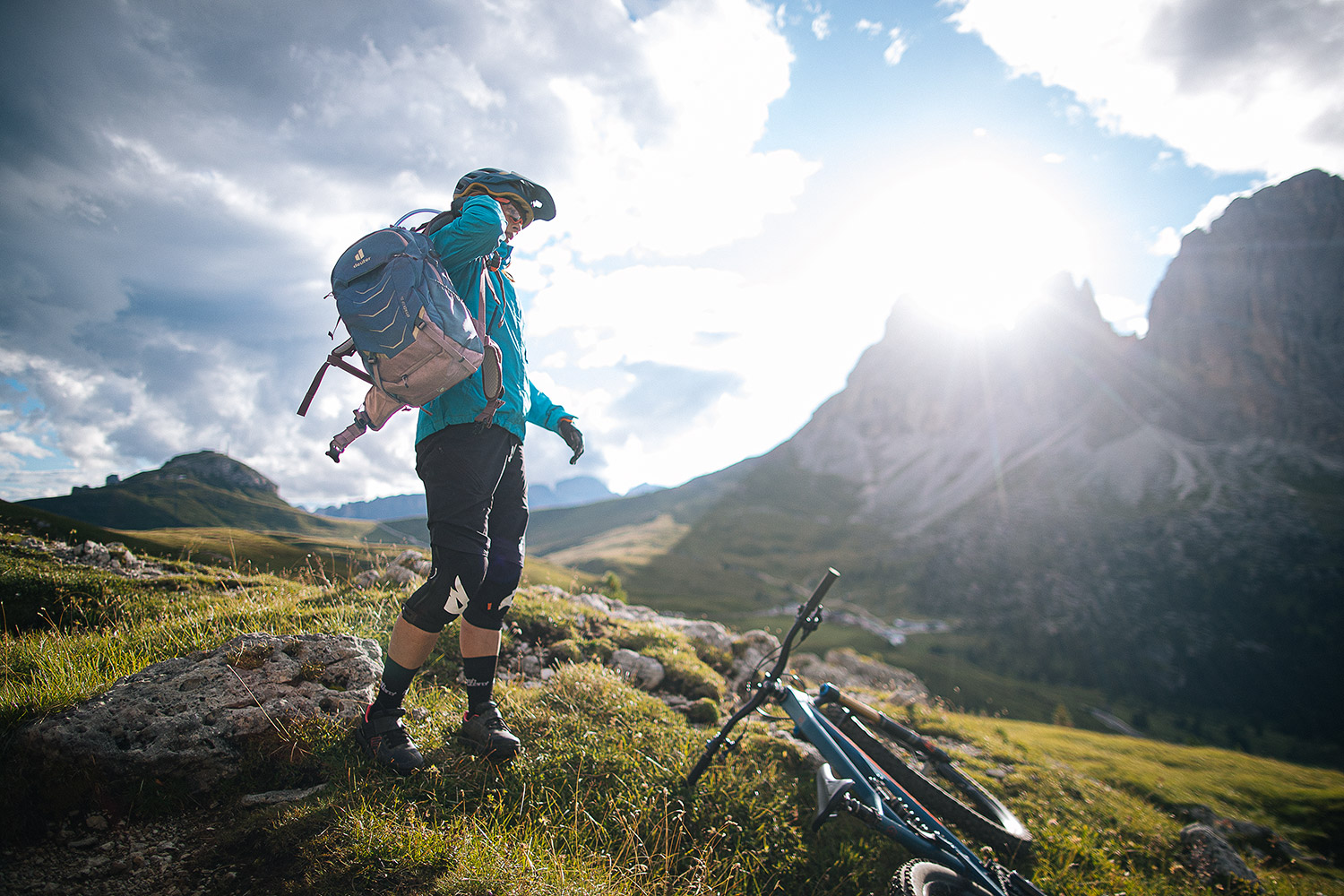 Bild Eine Frau in Fahrradklamotten steht in alpiner Landschaft und zieht ihren Fahrradrucksack auf.