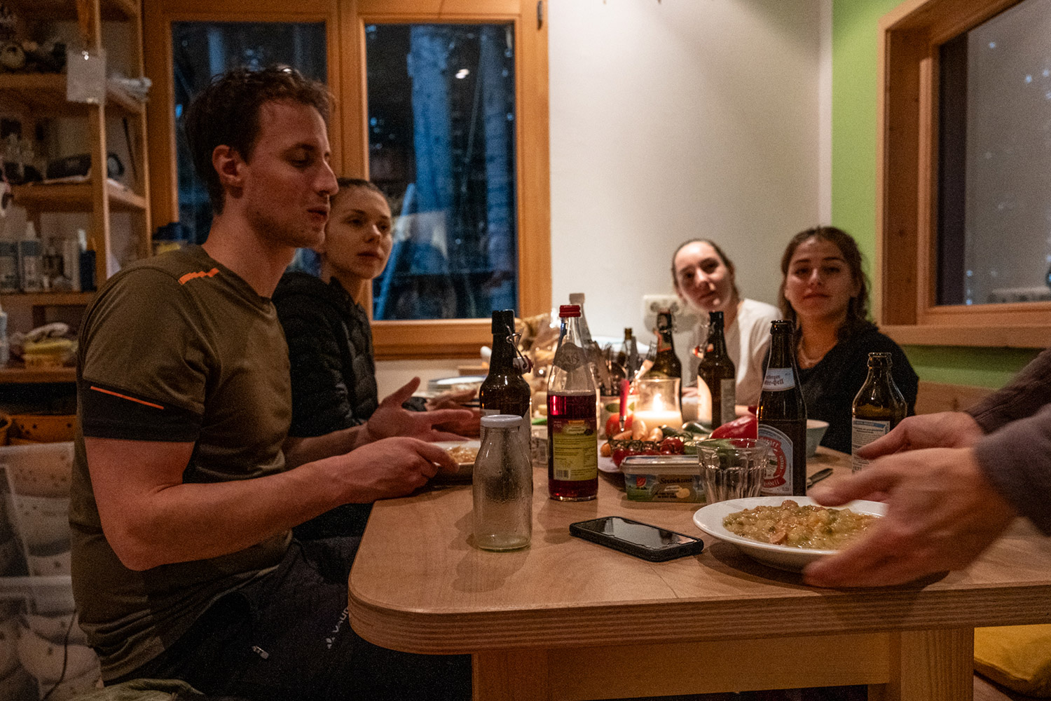Bild Vier Menschen sitzen auf einer Hütte beim Essen, eine weitere Person stellt einen Teller Eintopf auf den Tisch.