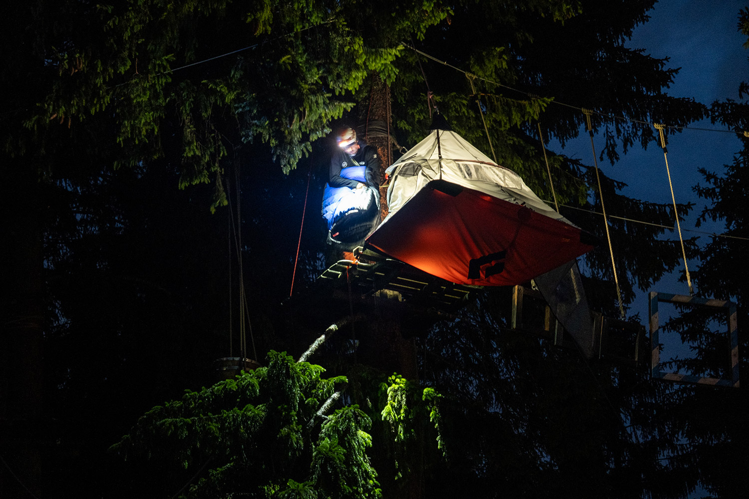 Bild Ein Mann steht mit Schlafsack in der Hand im Dunkeln auf einer Plattform neben der Portaledge. Er beleuchtet alles mit einer Stirnlampe.