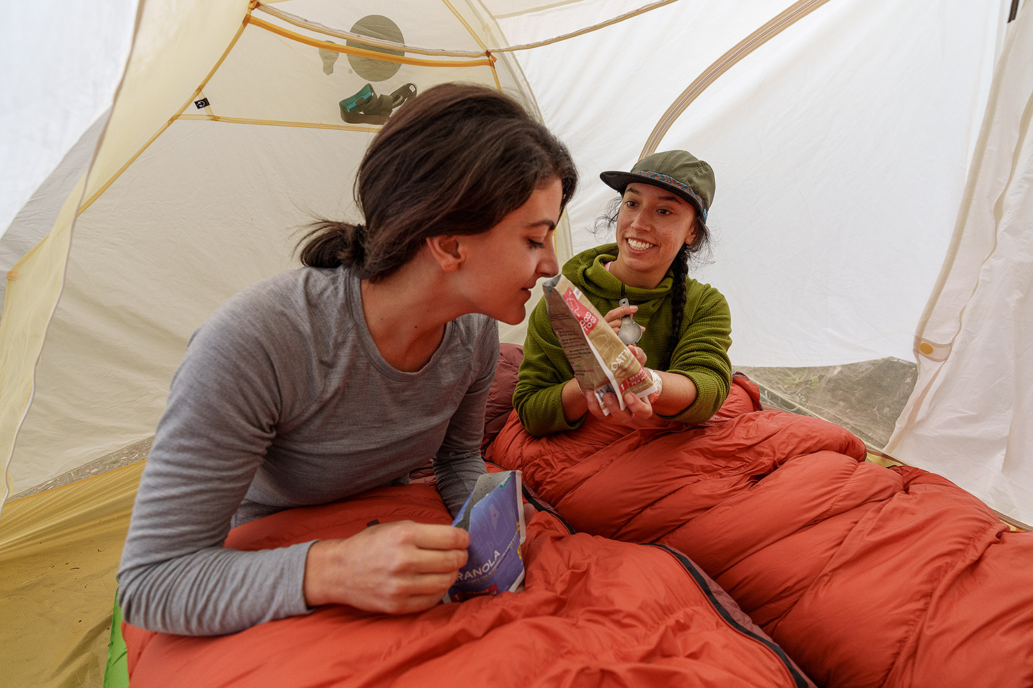 Bild Zwei Frauen sitzen in einem Zelt in ihren roten Schlafsäcken und bereiten sich Tüten mit Trekkingnahrung zu.