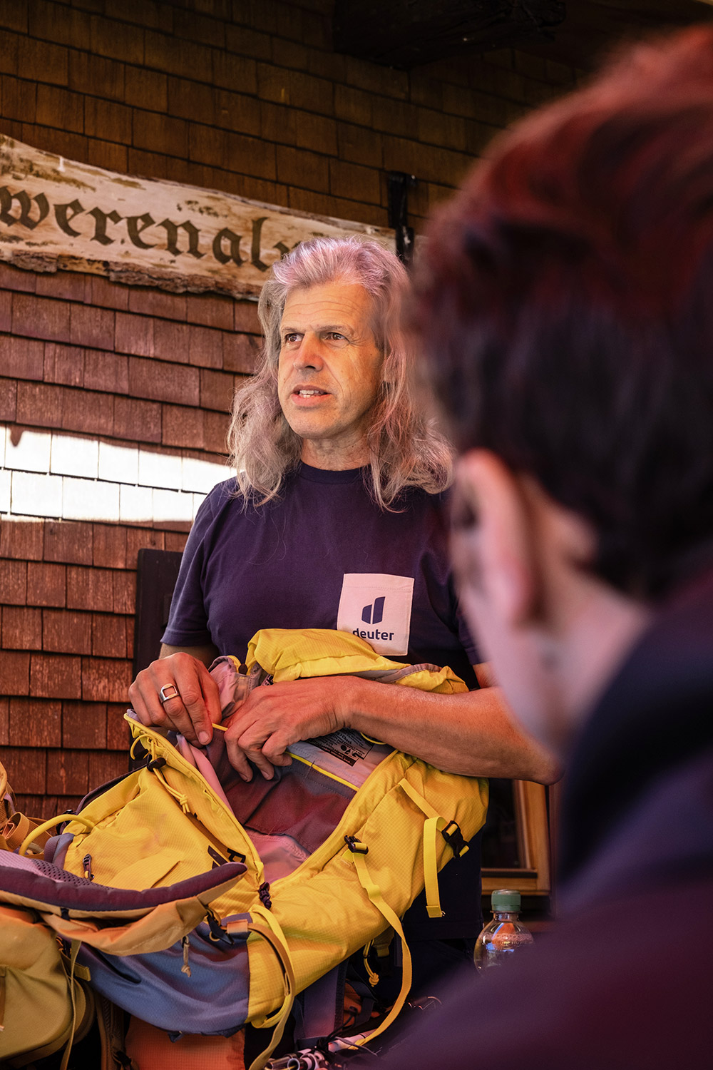 Bild Ein Mann mit langen, grauen Haaren erklärt etwas an einem Rucksack. Im Vordergrund sieht man den Zuhörer.