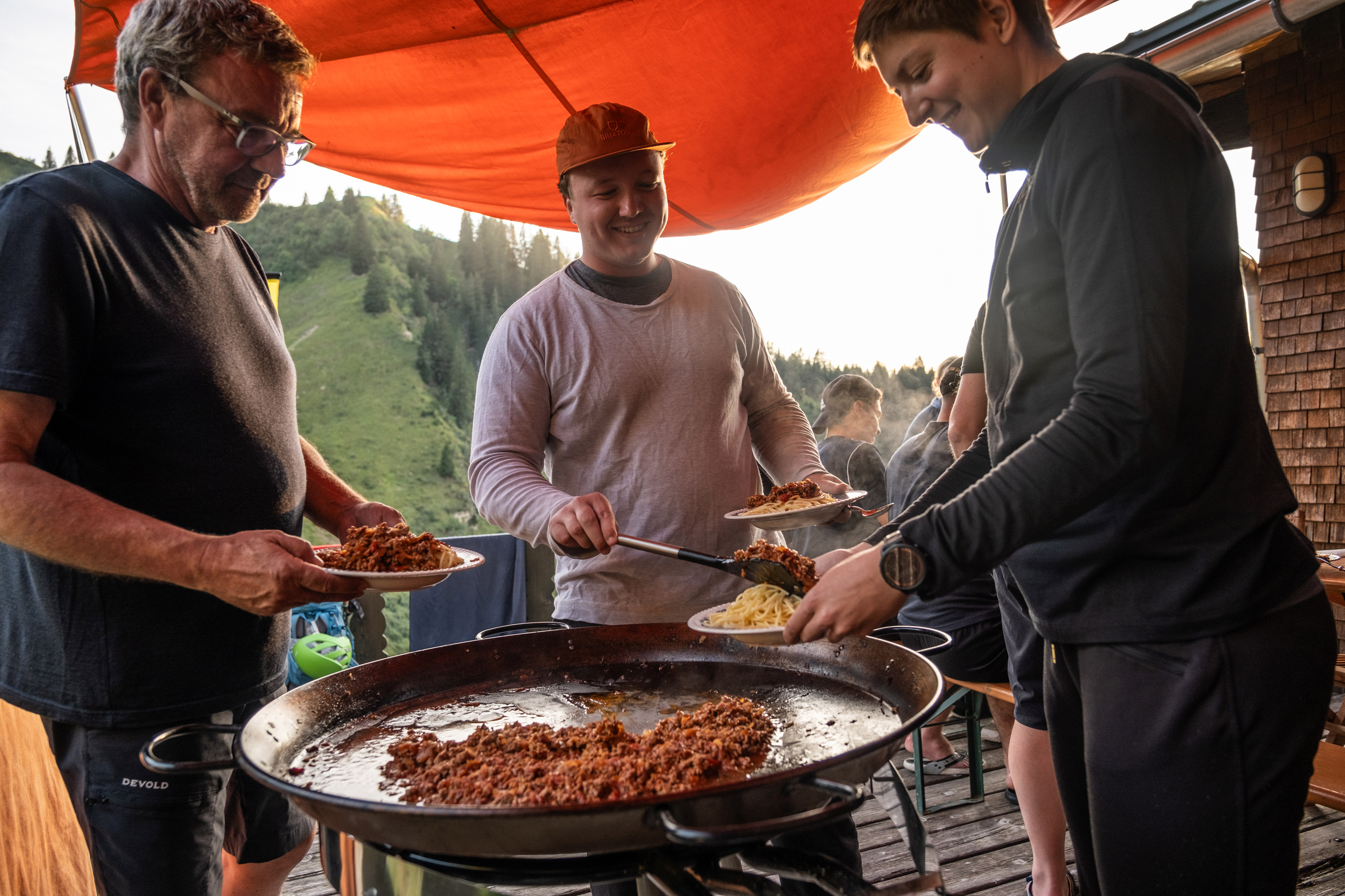 Bild 3 Personen stehen an einer großen Pfanne und schöpfen sich Bolognese Sauce. 