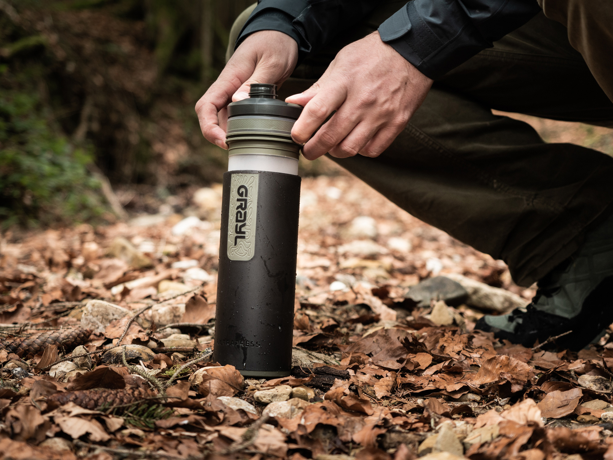 Bild Eine Grayl-Filterflasche steht auf belaubtem Waldboden und das Filterelement wird hineingedrückt.