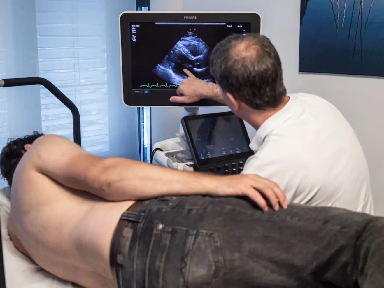 Bild Mann liegt auf einer Liege beim kardiologischen Check-Up. Arzt erklärt etwas am Monitor.