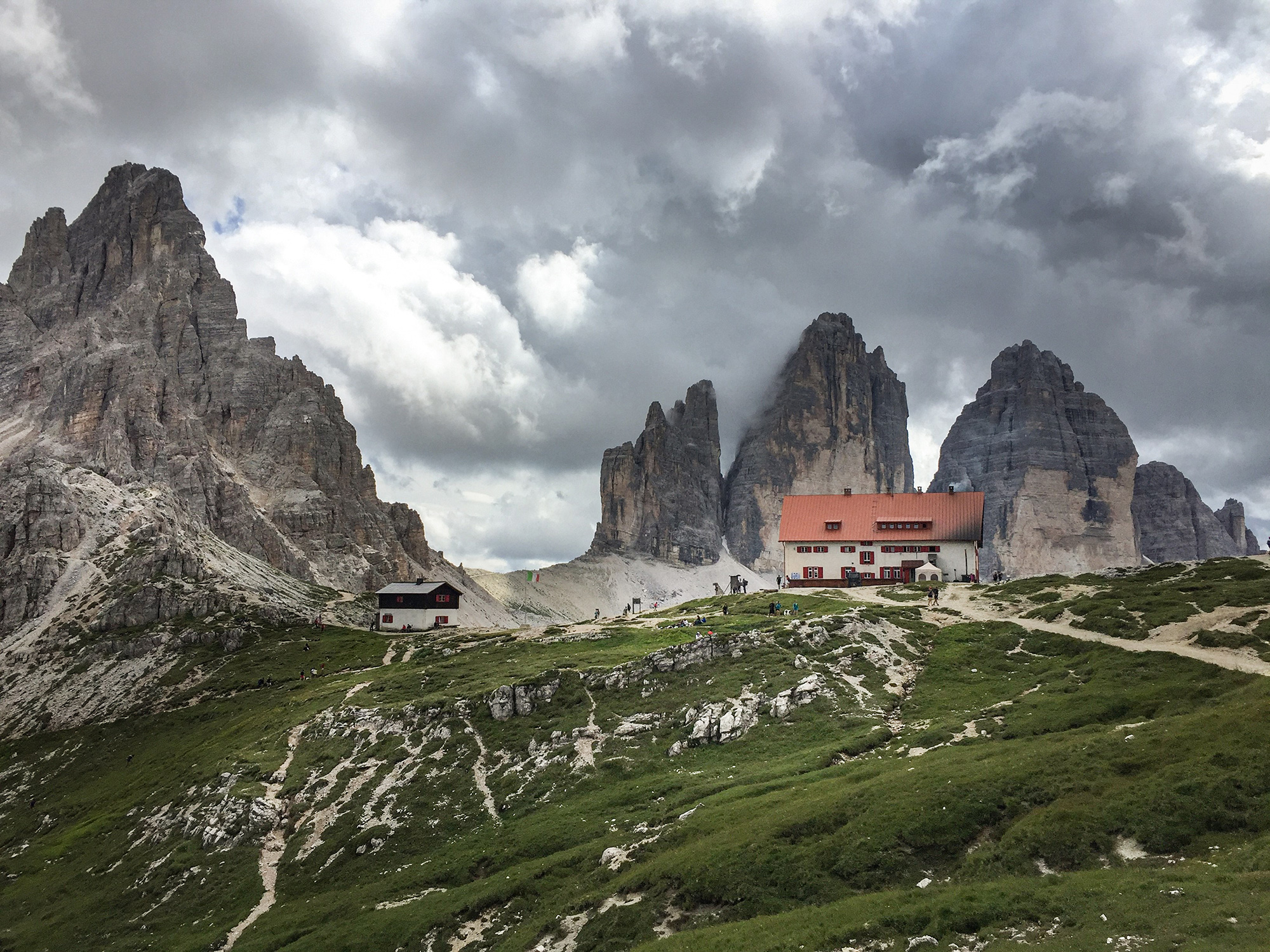 Bild Ein bewölkter Himmel. Grünes Gras mit einem Schotterweg, der zu einer Hütte mit rotem Dach führt. Im Hintergrund sind 4 Gipfelspitzen zu sehen, die steil in den Himmel ragen.