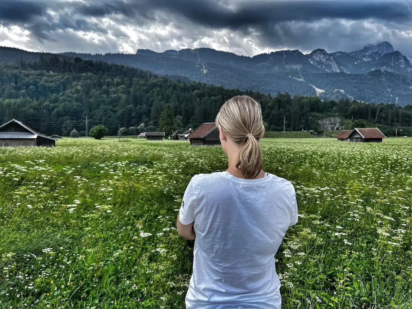 Bild Frau blickt über eine Bergwiese voller Almhütten in Richtung dunkler Wolken