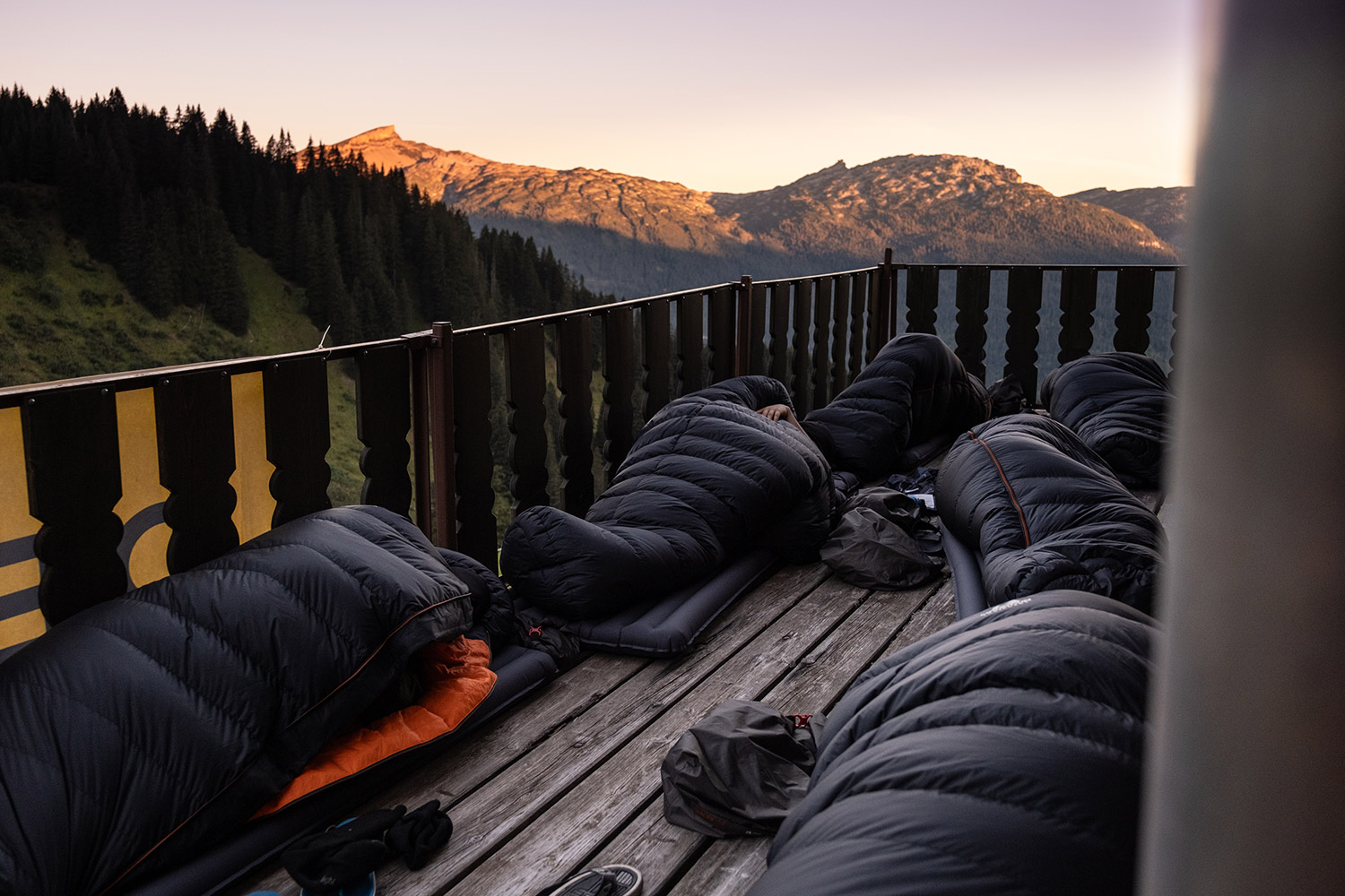 Bild Viele Personen liegen in Schlafsäcken auf einer Terrasse, im Hintergrund sieht man die Sonne über einem Berg aufgehen.