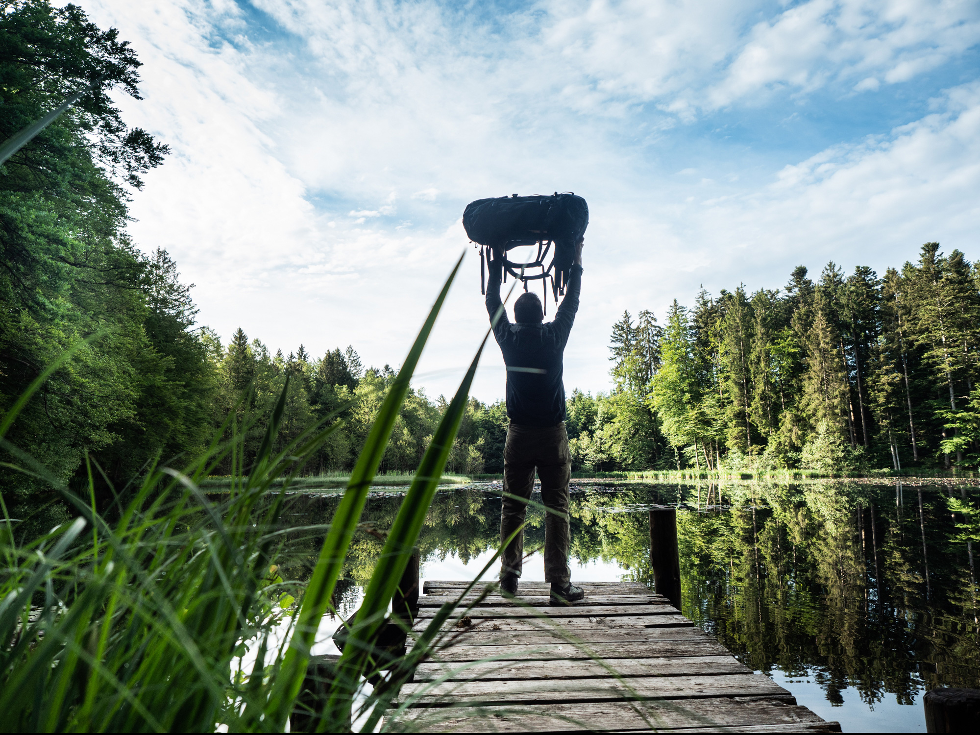 Bild Mann steht auf einem Steg am See und streckt seinen Rucksack in die Luft