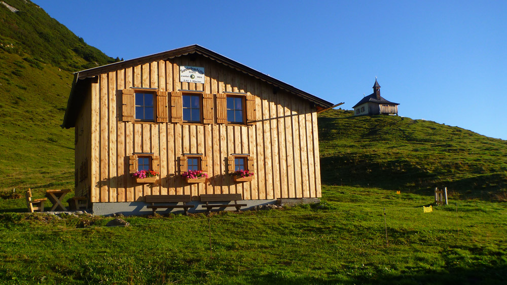 Bild Berghuette mit Kapelle im Hintergrund im warmen Abendlicht