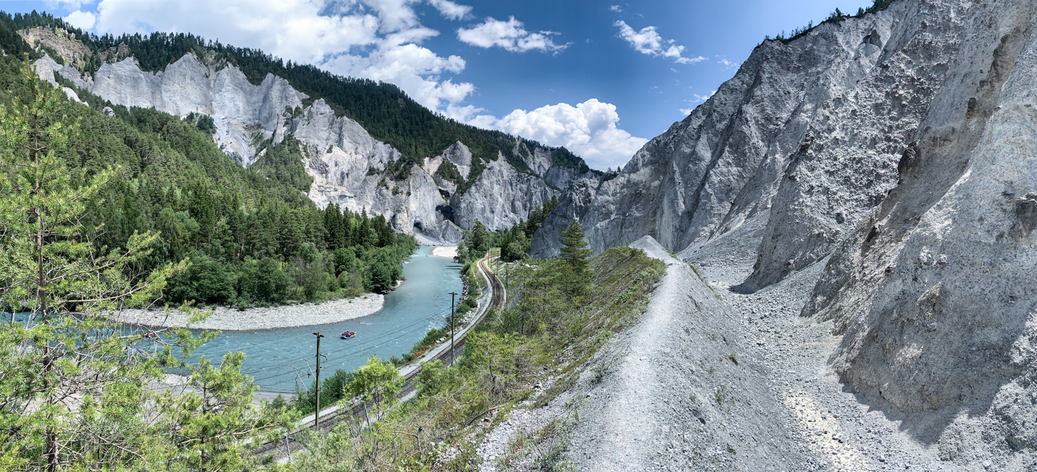 Bild Blick in die Rheinschlucht, auf dem Fluss sieht man ein Boot. Daneben einen Wanderweg und etwas tiefer Gleise.